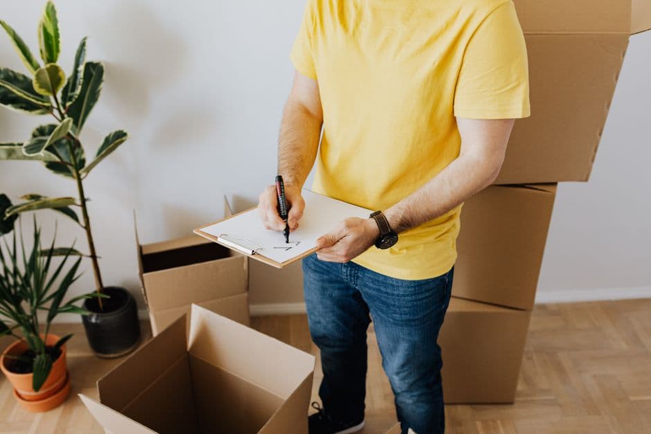 From above crop anonymous male wearing casual outfit ticking important notes in clipboard while packing carton boxes during moving out